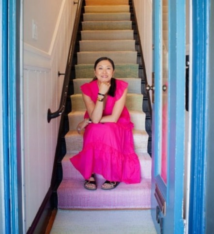 Woman with black hair in ponytail wearing a fuchsia dress smiling sitting on a steep multicolor–carpeted staircase